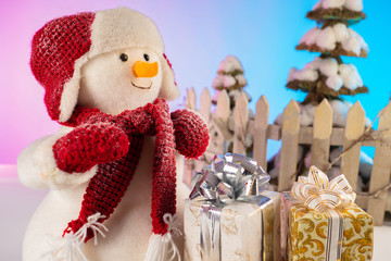 Congratulations to the New year. Plush snowman with gifts. Preparation for Christmas. Greeting card. Christmas decor. Packaging of Christmas gifts. Toy snowman with a red hat, scarf and mittens.
