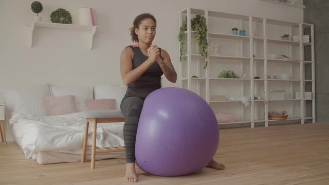 Determined motivated stunning african american woman in sports clothes working out with fitball, doing seated stability ball adductor squeeze exercise in domestic room during intense fitness training.