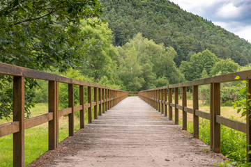 Obraz premium Wooden bridge over a field with meadow and wooded hills