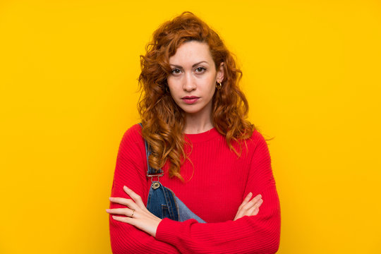 Redhead woman with overalls over isolated yellow wall keeping arms crossed