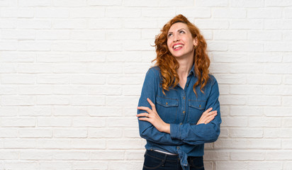 Redhead woman over white brick wall looking up while smiling