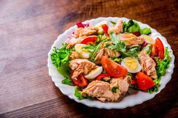 Fresh salad with fish, arugula, eggs,red pepper, lettuce, fresh sald leaves and tomato on a white plate on wooden table background