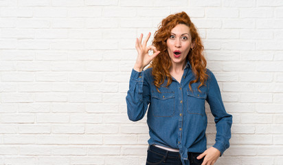 Redhead woman over white brick wall surprised and showing ok sign