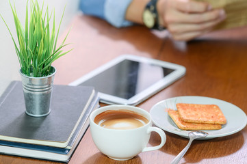 White cup of coffee on wooden table with blurred image of man reading news paper.