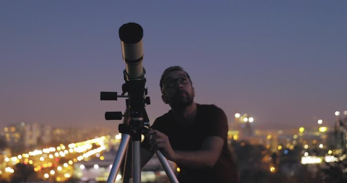 Astronomer with a telescope watching at the stars and Moon with blurred city lights in the background.