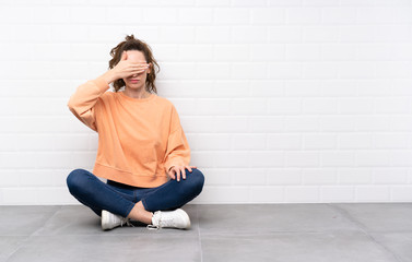 Young woman with curly hair sitting on the floor covering eyes by hands
