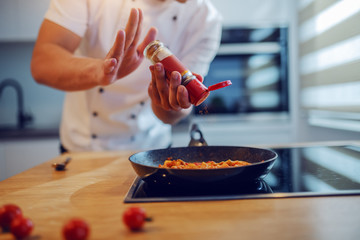 Close up of Caucasian chef adding chilli powder into tagliatelle on stove. Kitchen interior.