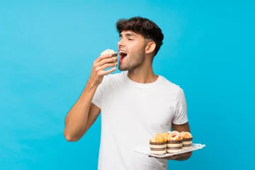 Fotobehang Young handsome man over isolated blue background holding mini cakes and eating it © luismolinero