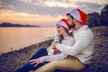 Handsome caucasian couple in love sitting in coast near river. Man hugging woman and kissing her in the head while woman looking at beautiful view. Both having santa hats on heads.