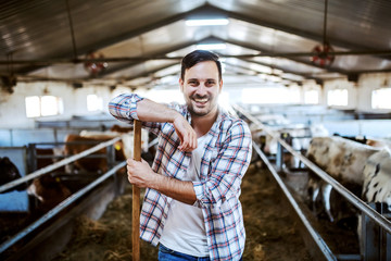 Handsome smiling caucasian farmer leaning on hay fork and looking at camera. In background are...