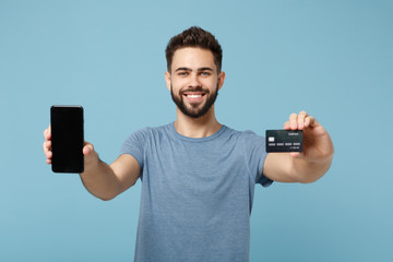 Young smiling man in casual clothes posing isolated on blue wall background, studio portrait. People lifestyle concept. Mock up copy space. Hold mobile phone with empty blank screen, credit bank card.
