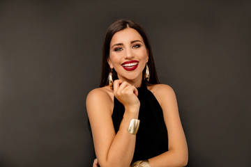 Portrait of a cheerful beautiful girl wearing black dress and gold earning standing over isolated black background