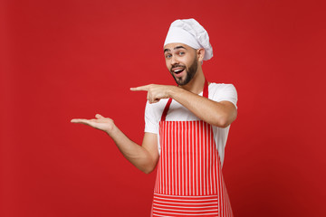 Cheerful young male chef cook baker man in striped apron white t-shirt toque chefs hat posing isolated on red background. Cooking food concept. Mock up copy space. Pointing index finger, hand aside.