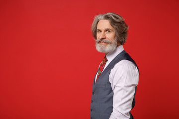 Side view of elderly gray-haired mustache bearded man in classic shirt vest colorful tie isolated on red wall background studio portrait. People lifestyle concept. Mock up copy space. Looking camera.