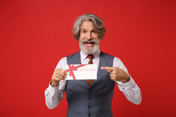Cheerful elderly gray-haired mustache bearded man in classic shirt vest and colorful tie isolated on red background. People lifestyle concept. Mock up copy space. Pointing finger on gift certificate.