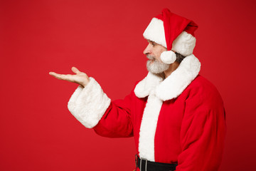 Fototapeta na wymiar Side view of elderly gray-haired mustache bearded Santa man in Christmas hat posing isolated on red background. New Year 2020 celebration holiday concept. Mock up copy space. Hold something in hand.