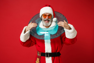 Fototapeta na wymiar Excited elderly gray-haired mustache bearded Santa man in Christmas hat sunglasses posing isolated on red background. New Year 2020 celebration concept. Mock up copy space. Holding inflatable ring.