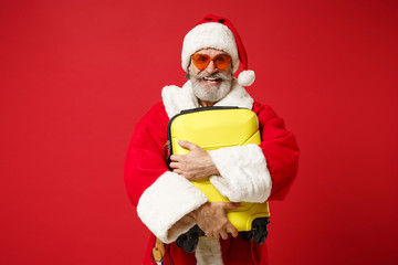 Joyful elderly gray-haired mustache bearded Santa man in Christmas hat, sunglasses posing isolated on red background. New Year 2020 celebration holiday concept. Mock up copy space. Holding suitcase.