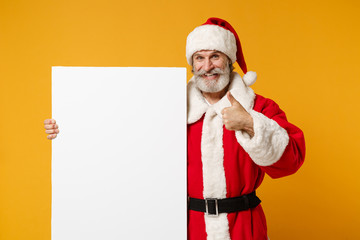 Fototapeta na wymiar Elderly gray-haired mustache bearded Santa man in Christmas hat isolated on yellow background. New Year 2020 celebration concept. Mock up copy space. Holding white empty billboard, showing thumb up.