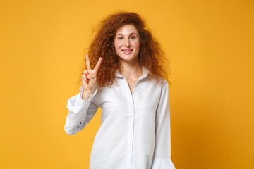 Smiling young redhead woman girl in casual white shirt posing isolated on yellow orange wall background in studio. People sincere emotions lifestyle concept. Mock up copy space. Showing victory sign.