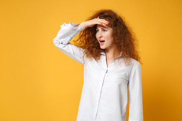 Young redhead woman girl in casual white shirt posing isolated on yellow orange background in studio. People lifestyle concept. Mock up copy space. Holding hand at forehead looking far away distance.