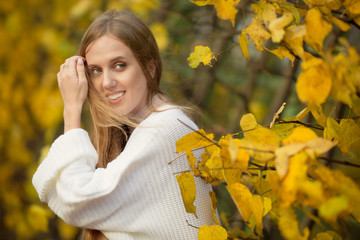 Portrait of a happy beautiful cute girl among yellow foliage. A smiling attractive young woman in white clothes looks away and straightens her blond hair with her hand. Autumn mood. Fall season.