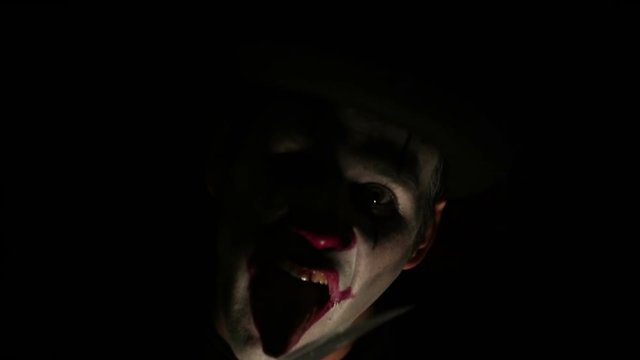 Terrible clown threatens his victim with a knife.Terrible man in the makeup of a clown looks at the camera, laughs and licks a knife blade. The terrible grimaces of a clown looking at the camera. Hall
