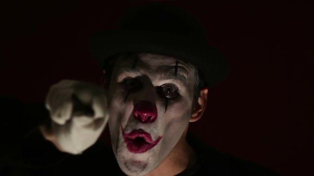 Terrible clown threatens his victim with a knife.Terrible man in the makeup of a clown looks at the camera, laughs and licks a knife blade. The terrible grimaces of a clown looking at the camera. Hall