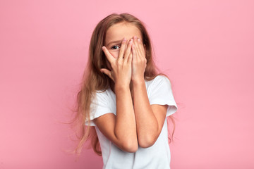 emotional little girl covers her face with her hand isolated over pink background, child watching...