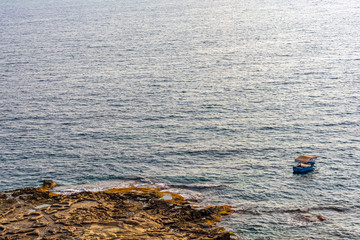 Lonely boat floating on the Mediterranean sea near Sliema coastline, Malta. Fishing boat is moored in the open sea near the Sliema shore and sandstone beach in the warm summer evening.