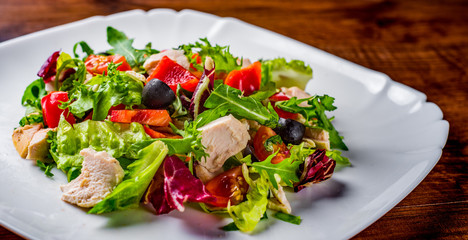 Fresh salad with chicken breast, arugula, black olives,red pepper, lettuce, fresh sald leaves and tomato on a white plate on wooden table background