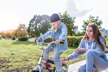 Mom woman teaches riding bike, little boy son 3-5 years old, training rides, in summer city, in autumn in park, casual clothes. Caring help and support in learning first lessons, balance.