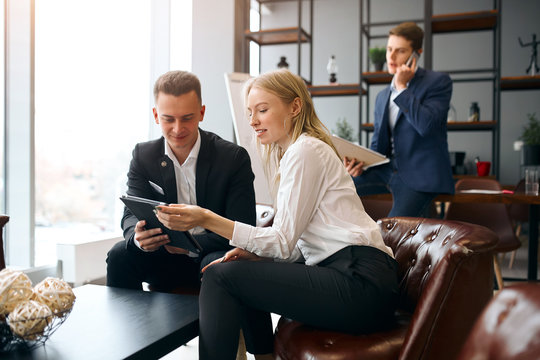 blonde woman recommends client to cooperate with their company, showing , advertsing goods while sitting on the armchair, mman talking with smartphone on the background of the photo