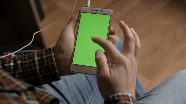 A man connects a headset to a smartphone to listen to music and watch movies. Male using smartphone with green screen while sitting on in room.