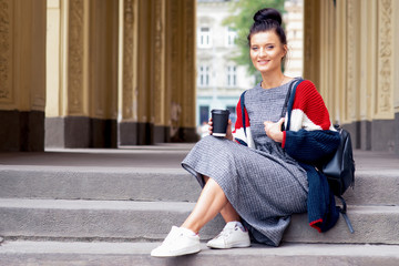 Adult student girl is holding paper coffee cup on stairs. Smiling student woman holds coffee cup on the university stairs.