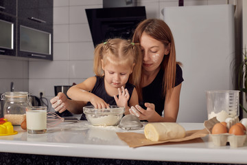 Obraz na płótnie Canvas Mother and daughter preparing a sweet cake using flour, milk, sitting on chairs at a table in a modern kitchen. Girl holding a whisk, stirring eggs in a bowl, preparing pancake dough with her mom.
