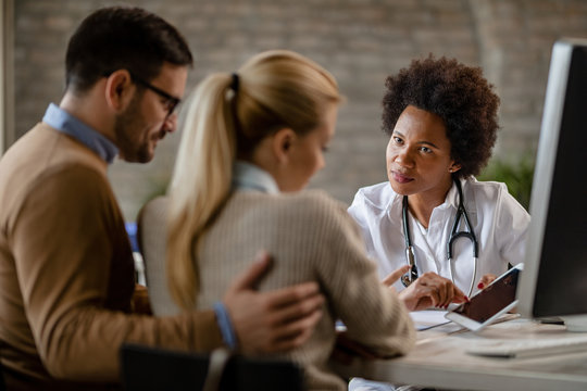 African American Female Doctor Using Touchpad While Talking To A Couple At Clinic.