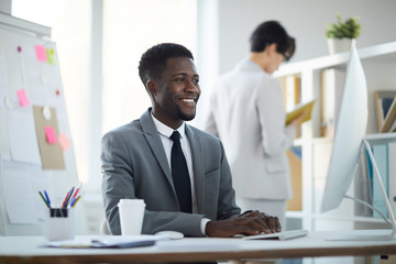 Portrait of young African-American businessman smiling happily while using computer sitting at workplace in office, copy space