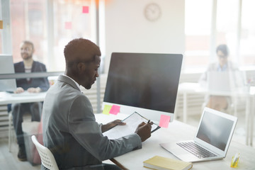 Side view portrait of African-American office worker sitting at workplace and filling forms, shot from behind glass, copy space