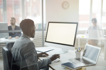 Back view portrait of African-American office worker using computer with black screen while sitting at workplace, shot from behind glass, copy space