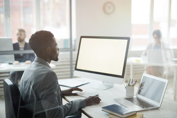 Side view portrait of African-American office worker using computer with black screen while sitting at workplace, shot from behind glass, copy space