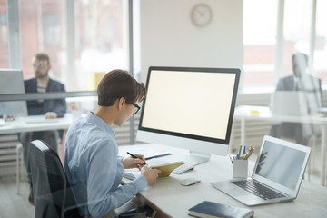 Back view portrait of young office worker taking notes while sitting at workplace, shot from behind glass, copy space