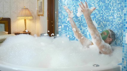 A young man with a green mask on his face and in a shower cap takes a bath with foam. the guy is dancing in the bathroom, his body is covered with thick white foam. copy space