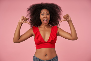 Joyous pretty curly lady with dark skin having belly button piercing pointing on herself with raised thumbs, looking at camera joyfully with wide mouth opened, posing over pink background