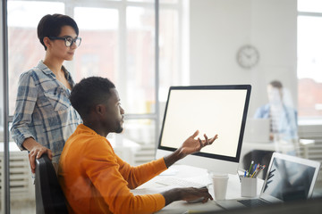 Side view portrait of young African-American man looking at blank computer screen and gesturing while discussing IT project with female colleague, copy space