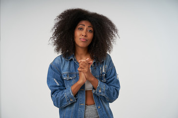 Portrait of young dark skinned lady with curly hair raising folded hands and looking at camera plaintively, pursing lips and wrinkling forehead while posing over white background
