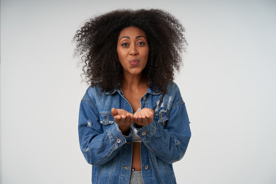 Portrait of charming young dark skinned woman with curly hair raising opened palms and blowing air kiss at camera, being in nice mood, standing over white background in casual clothes