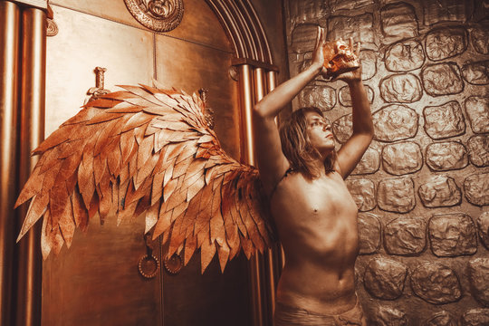 halloween make up. boy ready for party. man is covered in gold paint and poses with large angel wings and a skull in his hands against the backdrop of large doors in the Studio