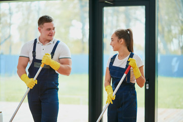 Staff of cleaning service work together, washing floor with mop. Wearing special clothes for cleaning. Panoramic window background