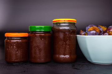 Jars with plum jam on a black background. In the background bowls with plums